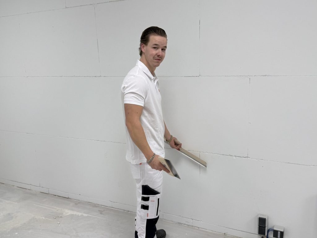 Stukadoor in wit werkpak houdt een lange stucrei tegen een kale wand en kijkt over zijn schouder de camera in