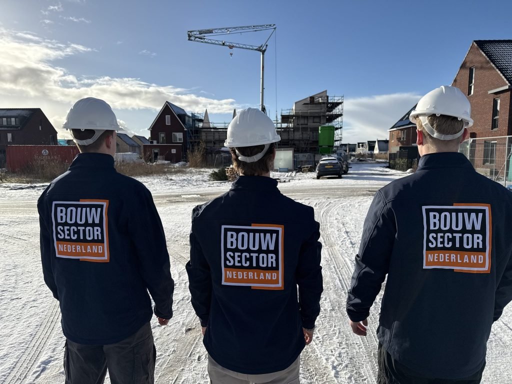 Drie vakmensen van Bouwsector Nederland met witte helm lopen over een besneeuwde bouwplaats met steigers en een kraan